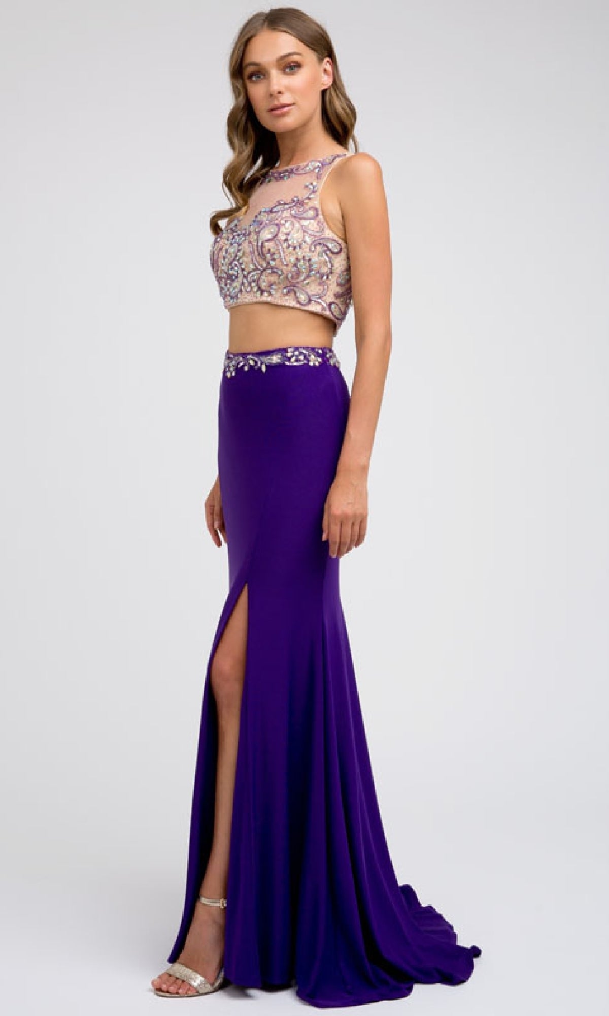 A woman models the Juliet 595 long prom dress—a purple two-piece with a beaded top and a long skirt featuring a side leg slit—paired with heels against a plain backdrop.